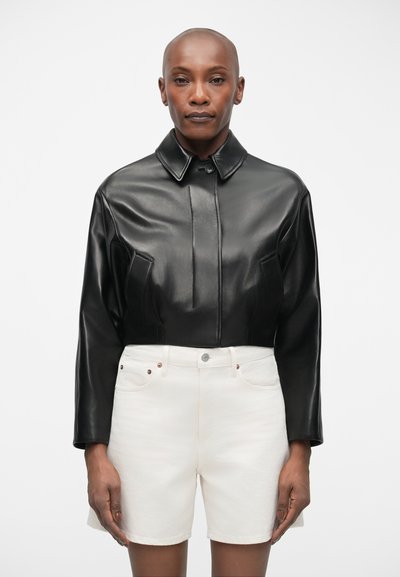 Veste en cuir noire cropped avec un col, présentant des détails de couture à l'avant, associée à des shorts en denim blancs. Design épuré et texture lisse.