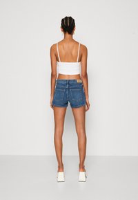 Žena s copánky, která má na sobě bílý crop top, modré džíny a bílé boty na podpatku, stojící zády k objektivu na jednobarevném pozadí.