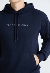 Sweat à capuche bleu marine en coton avec une poche avant, arborant le texte "TOMMY HILFIGER" en broderie blanche et rouge sur la poitrine.