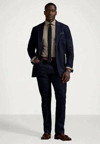 Vīrietis ar tumši zilu žaketi, pārbaudes krekli, tumši zilu kaklu saiti, tumšajiem biksēm un brūnām pusgludām kurpēm, stāv ar vienu roku kabatā.