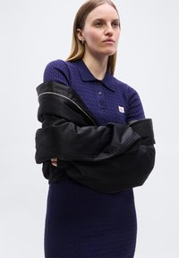 Γυναίκα με ίσια ξανθά μαλλιά φοράει μοβ πλεκτό φόρεμα πόλο με σχέδιο κοτσίδα και μαύρο σακάκι περασμένο στους ώμους της, σε απλό φόντο.