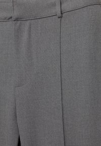 Gros plan sur un pantalon gris sur mesure montrant la taille, les passants de ceinture et le détail du pli avant sur un tissu texturé.