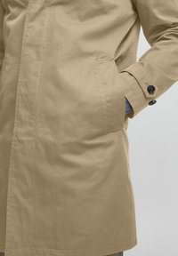Manteau beige à manches longues avec un poignet à bouton unique, poches latérales et un tissu lisse et texturé. Design simple et moderne.