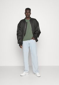 Giacca bomber nera su un maglione verde testurizzato, abbinata a jeans azzurri chiari e sneakers bianche. Outifit semplice e casual, con accenti minimi.