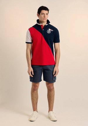 Hombre de pie vistiendo una camiseta tipo polo de colores rojo, azul marino y blanco, pantalones cortos cargo azul marino y zapatillas blancas contra un fondo liso.