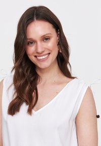 Biała, bezrękawnikowa bluzka z dekoltem w serek, z wiązaniami na ramionach i miękką, gładką fakturą. Udekorowana małymi brązowymi koralikami.