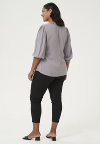 Blusa gris claro con mangas abullonadas y un escote con volantes, combinada con pantalones ajustados negros y sandalias de tacón, vista desde atrás.