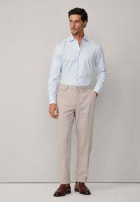 Camicia a righe azzurro chiaro con bottoni abbinata a pantaloni beige. Presenta un frontale piatto e tasche laterali. Le scarpe in pelle marrone completano l'outfit.