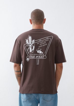 Homme portant un t-shirt marron avec un motif blanc de cactus, serpent enroulé, crâne de bœuf, et texte « Dans le pays du soleil et du sable, il était une fois dans l’Ouest. »