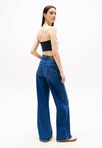 Wijduitlopende blauwe spijkerbroek heeft een hoge taille, achterzakken en een leren bruine patch. Het model draagt een cropped top en casual schoenen.