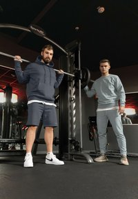 Twee mannen in gymkleding: de ene in een donkere hoodie en shorts, de andere in lichte sweatpants en een sweatshirt, staand bij een gewichtmachine.