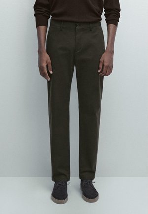 Dunkelgrüne Hose mit gerade geschnittener Passform, zwei seitlichen Taschen und einem vorderen Knopfverschluss, kombiniert mit dunklen Schuhen. Weicher Stoff. 