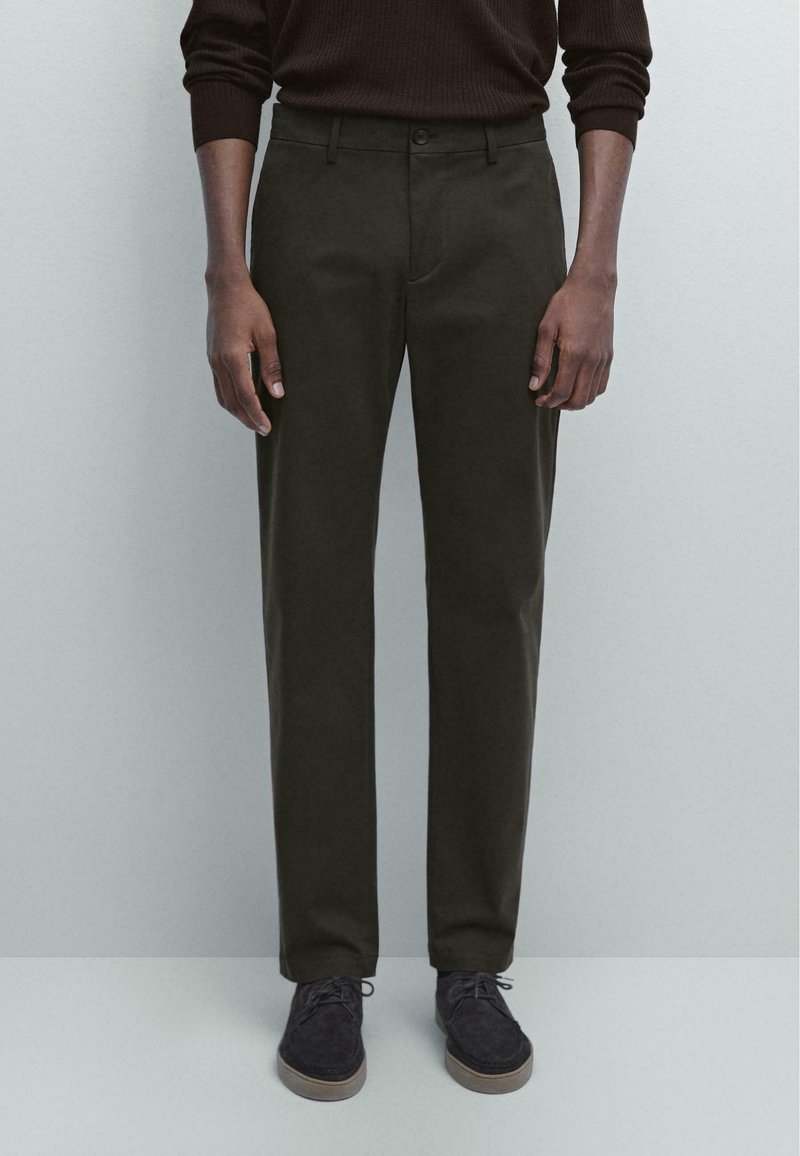 Dunkelgrüne Hose mit gerade geschnittener Passform, zwei seitlichen Taschen und einem vorderen Knopfverschluss, kombiniert mit dunklen Schuhen. Weicher Stoff.