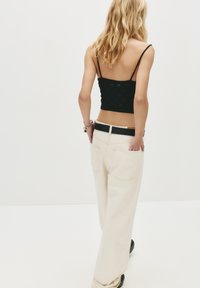 Een vrouw met blond haar draagt een zwarte cropped tanktop, beige wijde broek met een zwarte riem en een zilveren armband, terwijl ze tegen een witte achtergrond staat.