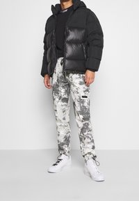Mann in schwarzer Pufferjacke, schwarzem Sweatshirt, grauen und weißen Batik-Cargohosen sowie weißen Sneakers mit schwarzen Schnürsenkeln vor einfarbigem Hintergrund.
