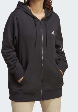Person iført en sort Adidas hættetrøje med lynlås, hætte og forlommer, hænderne i lommerne, kombineret med lysebeige bukser.