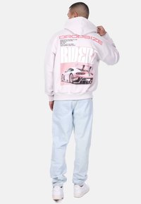 Weißer Hoodie mit einem großen rosa Grafik eines Autos auf dem Rücken, mit Text, der die Spezifikationen beschreibt; kombiniert mit hellblauen Jeans und weißen Sneakers.