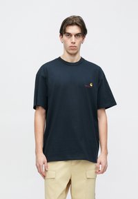 Σκούρο μπλε μπλουζάκι με στρογγυλή λαιμόκοψη και κοντά μανίκια, με ένα μικρό κίτρινο και κόκκινο λογότυπο στην αριστερή πλευρά του στήθους. Υλικό από βαμβάκι.