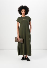 Olive green maxi dress with a round neck and short rolled sleeves, featuring a side slit. The model holds a brown quilted clutch. Black shoes.