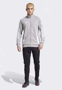 Szürke cipzáras sportdzseki fehér csíkokkal, fekete szűkített szárú nadrággal és fekete sportcipőkkel, narancssárga részletekkel.