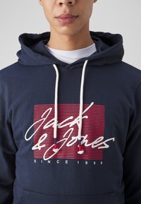 Felpa della marina con lacci bianchi, caratterizzata da una grafica a strisce rossa e bianca, "Jack & Jones," e il testo "SINCE 1990" nella parte inferiore.
