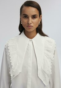 Blusa bianca con un grande colletto decorato con ruches in pizzo. Tessuto morbido, chiusura con bottoni e design a taglio dritto.