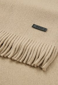 Beige neulehuivi, jossa on fringed-yksityiskohtia ja musta tuotetagi. Tekstuuri on sileä ja hieman joustava ulkonäöltään.