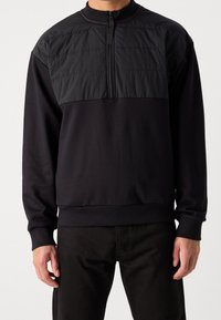 Sudadera negra con medio cierre frontal, que presenta un panel superior texturizado. Fabricada con material suave y con puños y cinturilla de canalé.