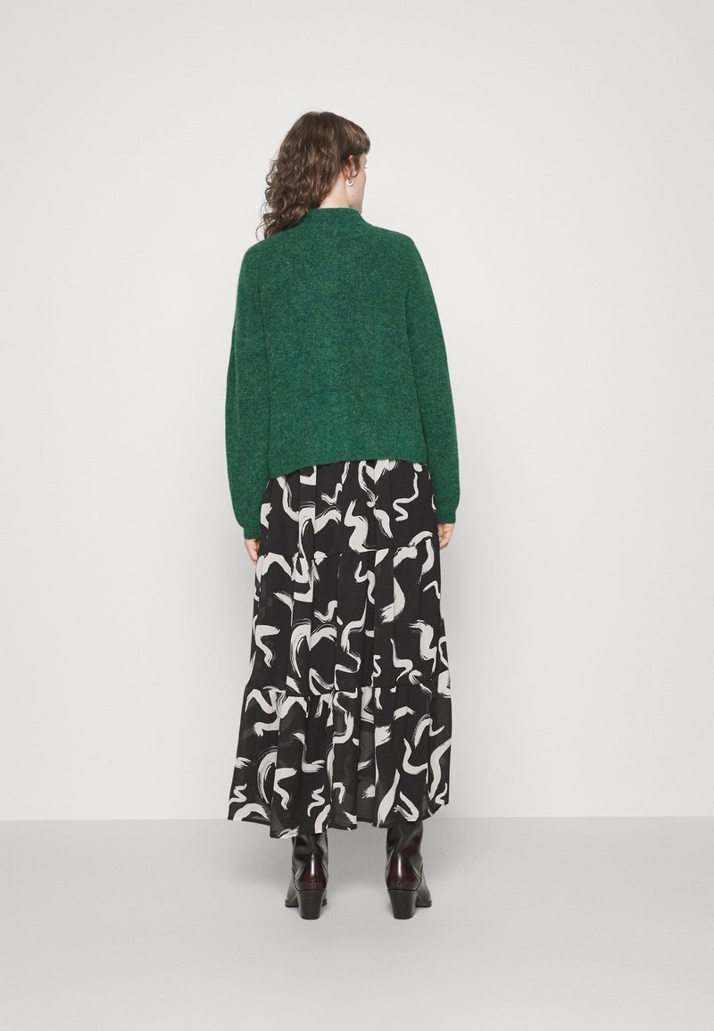 Pull vert texturé associé à une jupe maxi noire à motifs avec des designs abstraits blancs. Le modèle porte des bottes à talons sombres.