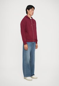 Sweat à zip maroon avec deux poches avant, associé à un jean bleu à jambes larges et des baskets blanches. Tissu lisse, coupe décontractée.