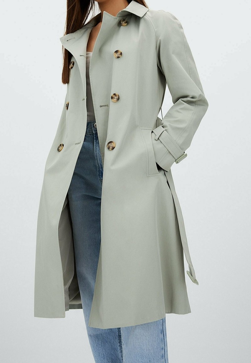Femme portant un long trench-coat gris clair avec de gros boutons et une ceinture, associée à un jean bleu et un haut blanc, debout avec une main dans la poche.