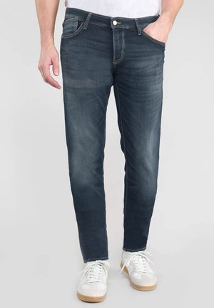Jeans en denim foncé avec une coupe slim, comportant des coutures visibles, cinq poches et une fermeture éclair, associés à des baskets blanches.