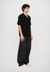 Cămașă neagră cu mâneci scurte și nasturi, cu un croi lejer, asortată cu pantaloni largi de culoare gri închis și adidași albi. Textură netedă.