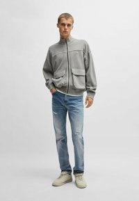 Ljusgrå zip-up jacka med två framfickor, bärs över en vit skjorta, tillsammans med ljusblå slitna jeans och beigefärgade sneakers.