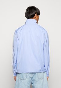 Lysblå jakke med høj krave, med hvide sidelapper og elastiske manchetter. Glat tekstur med svage lodrette linjer på stoffet.