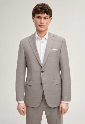 Giovane uomo con capelli ricci che indossa un abito grigio chiaro con una camicia bianca, in piedi davanti a uno sfondo neutro e semplice.