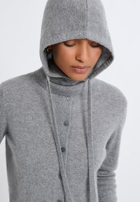 Cardigan gris en tricot avec capuche, doté d'un col montant, de cordons de serrage et de cinq boutons. Texture douce et coupe décontractée.