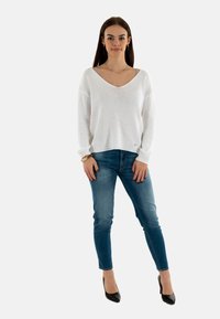 Suéter blanco de punto con escote en V, mangas largas y un dobladillo ligeramente corto, combinado con jeans ajustados azul y tacones altos negros.
