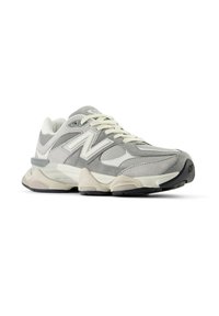 Sive in bele atletske superge New Balance z bež podplatom, mrežastimi in semiš pannami, vezalkami in logotipom na strani in jeziku, prikazane na beli podlagi.