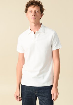 Giovane uomo con capelli ricci che indossa una polo bianca a maniche corte e jeans di denim scuro su uno sfondo beige uniforme.