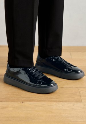 Zapatillas de charol negro con suela de goma texturizada, punta redonda y cordones negros a juego, combinadas con pantalones negros sobre un suelo de madera.