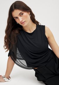 Sort ærmeløs top med draperet design og gennemsigtige sidepaneler. Glat tekstur med samlet detaljering ved skuldrene. Båret med sorte bukser.