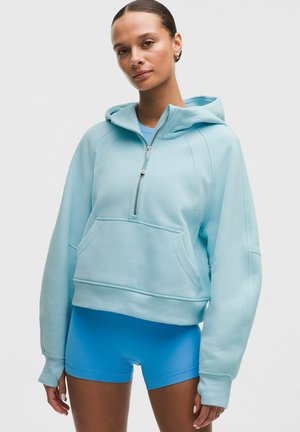 Kvinde iført en lyseblå cropped hoodie med forlommelukning og lynlås, kombineret med lyseblå tætsiddende shorts, stående mod en ensfarvet baggrund.