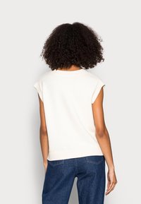 Cremefarbener ärmelloser Stricktop mit einem gerippten Halsausschnitt und Saum. Glatte Textur; lockere Passform, kombiniert mit blauen Jeans.