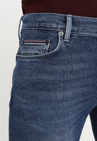 Tmavě modré džíny se středním přáním, s pětipočetným designem kapes, kovovým kováním a malým červeným a bílým akcentem na zadní kapse.