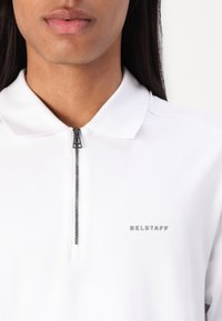 Balts polo krekls ar rāvējslēdzēja apkakli, ar minimālistisku dizainu un diskretu "BELSTAFF" logo kreisajā pusē. Gluda tekstūra.