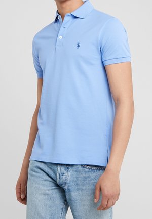 Mand iført en lyseblå kortærmet polo skjorte med krave og knapstolpe, kombineret med lyseblå denim jeans, står mod en ensfarvet baggrund.