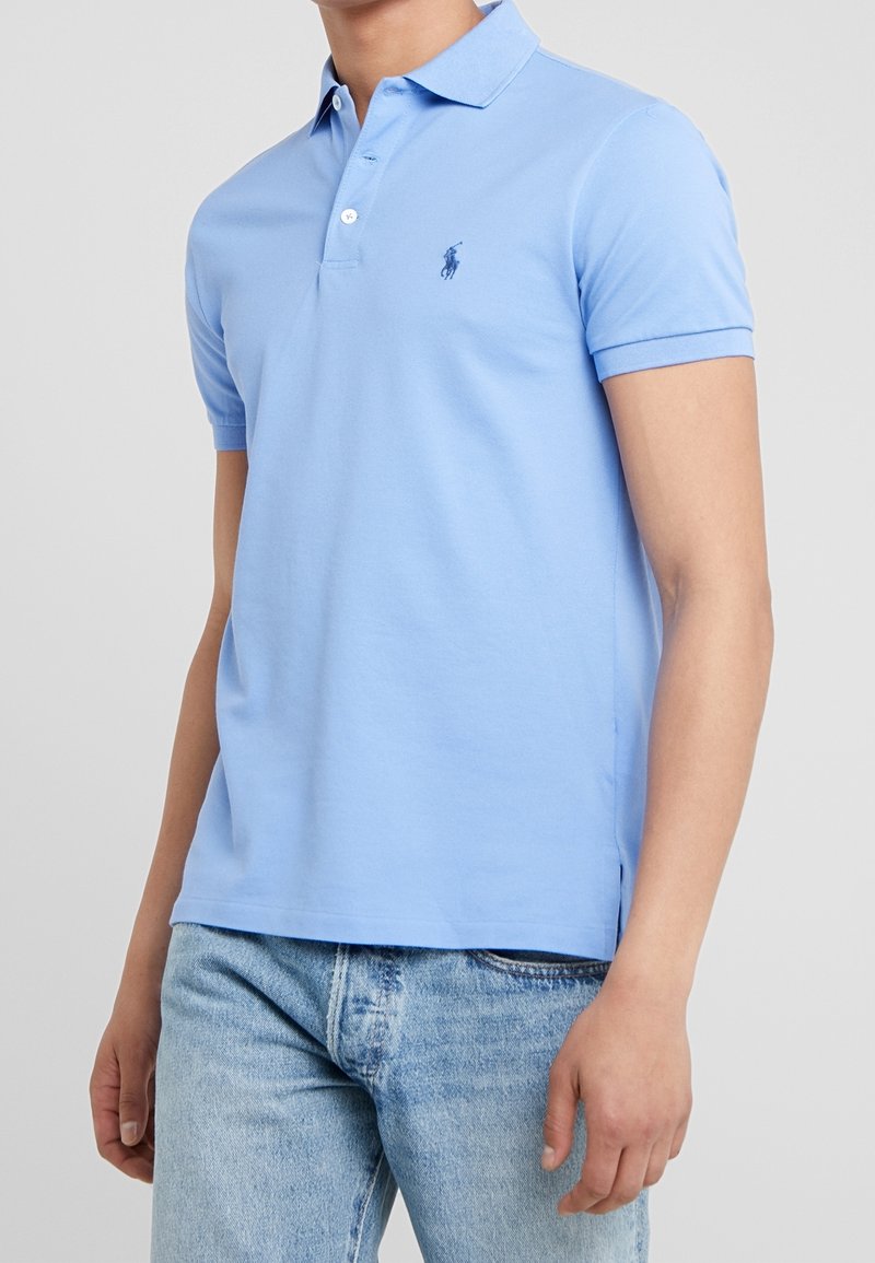 Homme portant un polo bleu clair à manches courtes avec col et patte de boutonnage, associé à un jean en denim bleu clair, debout devant un fond uni.