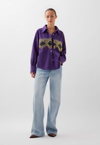 Camicia viola con bottoni, frange e design floreale ricamato sul petto, abbinata a jeans a vita alta azzurri e scarpe marroni.
