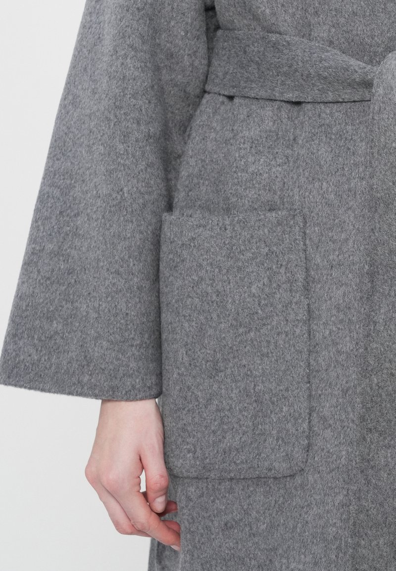 Ingrandimento della mano di una persona vicino a una grande tasca rettangolare sulla parte anteriore di un cappotto di lana grigia con maniche ampie.